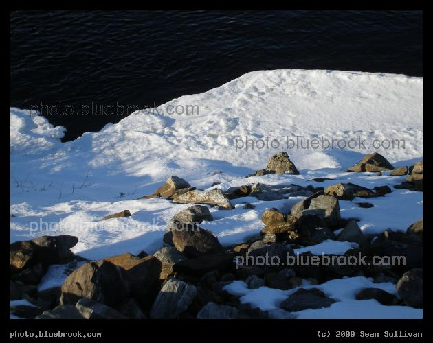Black and White Water - Looking down a rocky slope to the Androscoggin River in Lewiston ME, with dark liquid water meeting bright ice and snow.